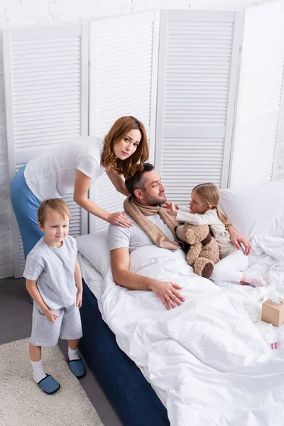 Vista de ángulo alto de la esposa y los niños que cuidan del padre enfermo en el dormitorio - foto de stock