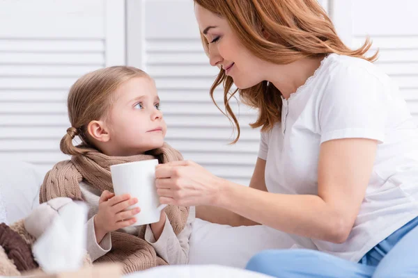 Madre cuidando de la hija enferma y dándole una taza de té en el dormitorio - foto de stock