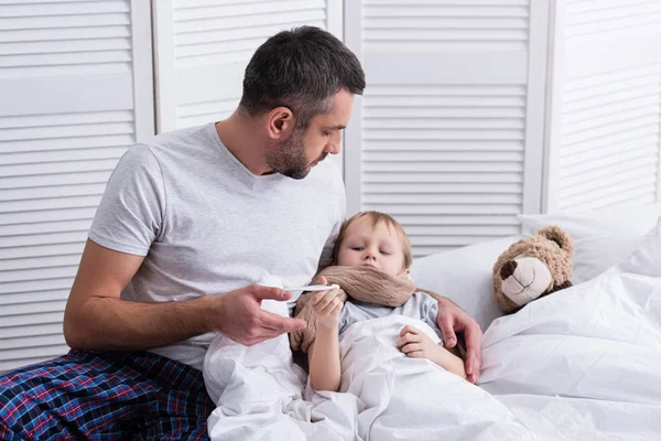 Padre cuidando al hijo enfermo en el dormitorio y comprobando su temperatura con termómetro - foto de stock