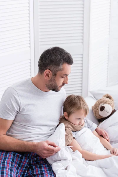 Handsome father taking care of sick son in bedroom and checking his temperature with thermometer — Stock Photo