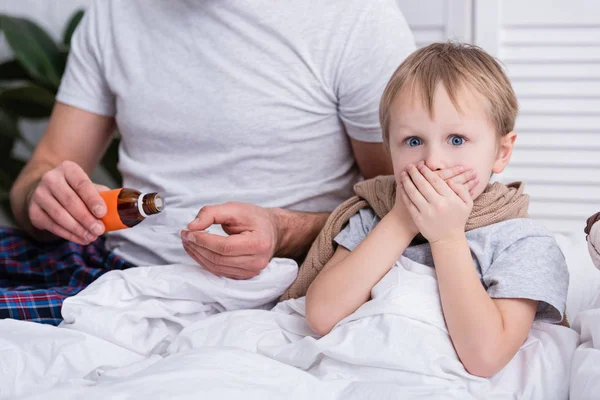 Cropped image of father pouring medicines in spoon for sick son, boy covering mouth and looking at camera — Stock Photo