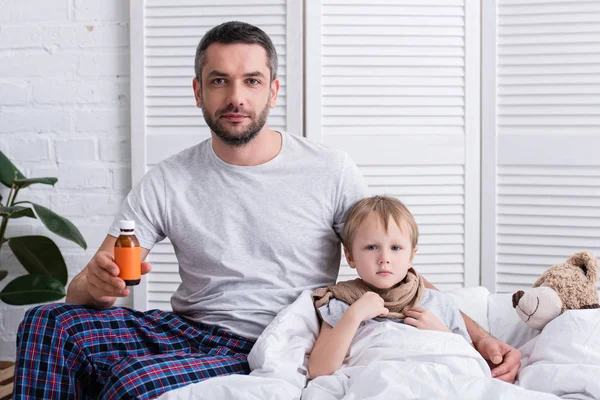 Père prendre soin de fils malade dans la chambre à coucher et montrant bouteille de médicaments — Photo de stock