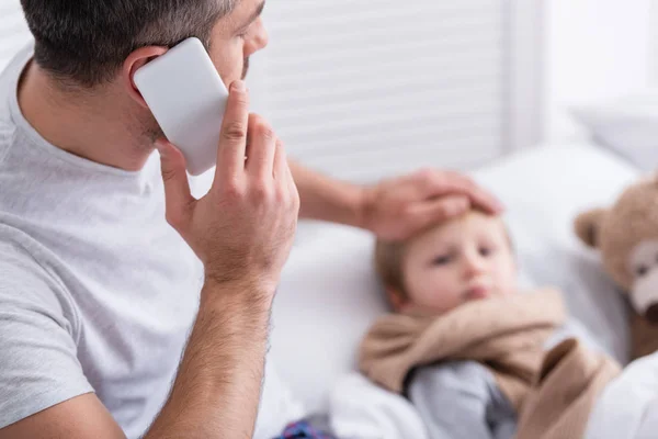 Enfoque selectivo del padre hablando por teléfono inteligente y tocando la frente del hijo enfermo en el dormitorio - foto de stock