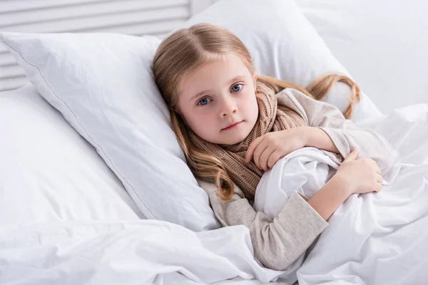 Enfant malade avec écharpe sur le cou couché dans le lit à la maison et regardant la caméra — Photo de stock
