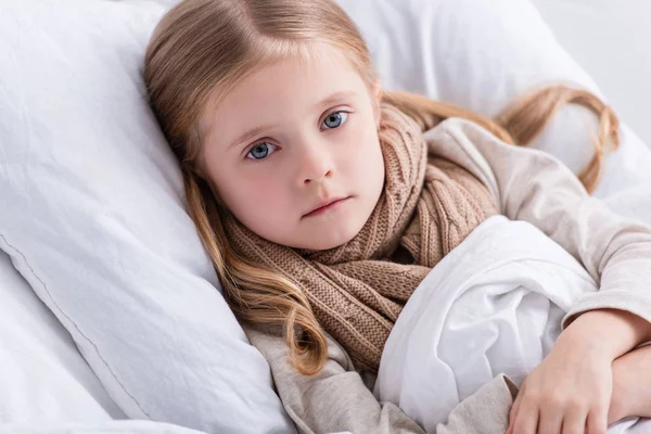 Retrato de niño enfermo con bufanda sobre el cuello acostado en la cama en casa y mirando a la cámara - foto de stock