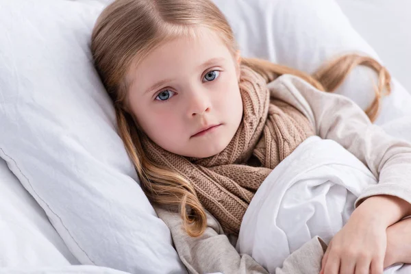 Retrato de niño enfermo con bufanda sobre el cuello acostado en la cama en casa y mirando a la cámara — Stock Photo