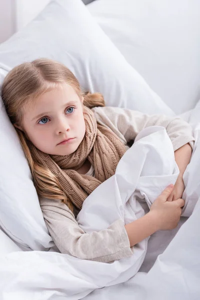 Niño enfermo con bufanda sobre el cuello acostado en la cama en casa y mirando hacia otro lado - foto de stock