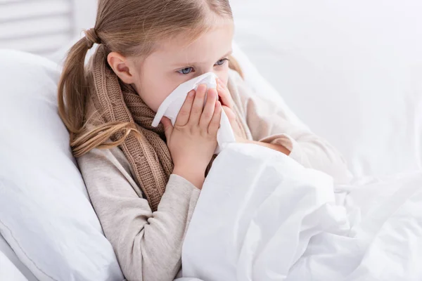 Enfant malade avec écharpe au-dessus du cou couché au lit et nez moucheté dans les tissus à la maison — Photo de stock