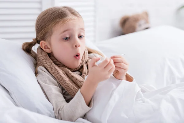 Criança doente chocado com cachecol sobre o pescoço deitado na cama e verificar a temperatura com termômetro em casa — Fotografia de Stock