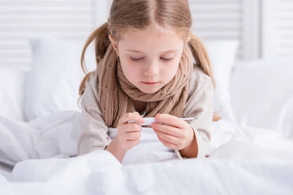 Krankes Kind mit Schal um den Hals liegt im Bett und schaut zu Hause auf Thermometer — Stockfoto