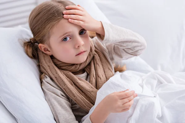 Krankes Kind mit Schal um den Hals im Bett liegend, Thermometer in der Hand und Stirn zu Hause angefasst — Stockfoto