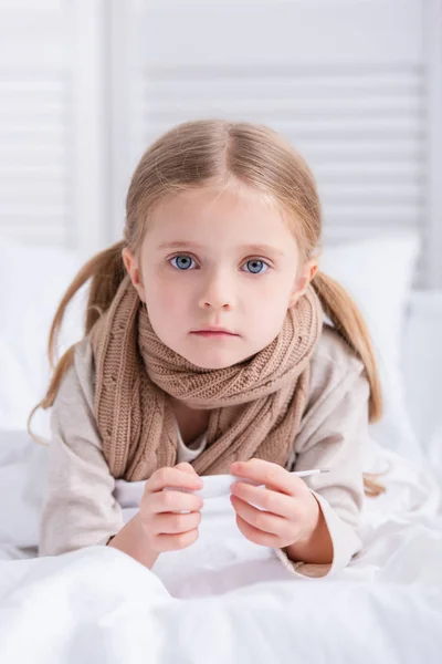 Больной ребенок с шарфом на шее лежит в постели, держит термометр и смотрит в камеру дома — стоковое фото