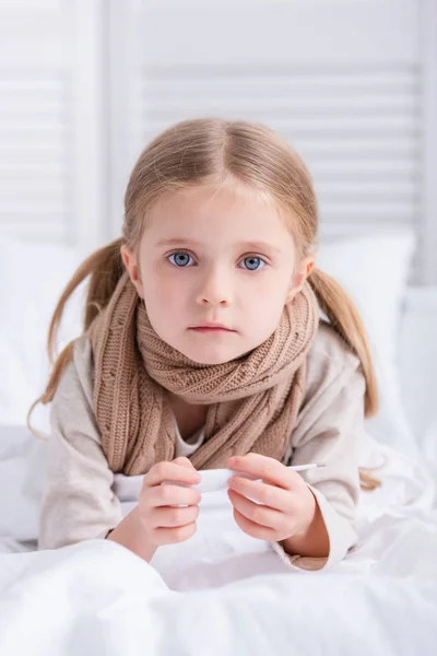 Niño enfermo con bufanda sobre el cuello acostado en la cama, sosteniendo el termómetro y mirando a la cámara en casa — Stock Photo