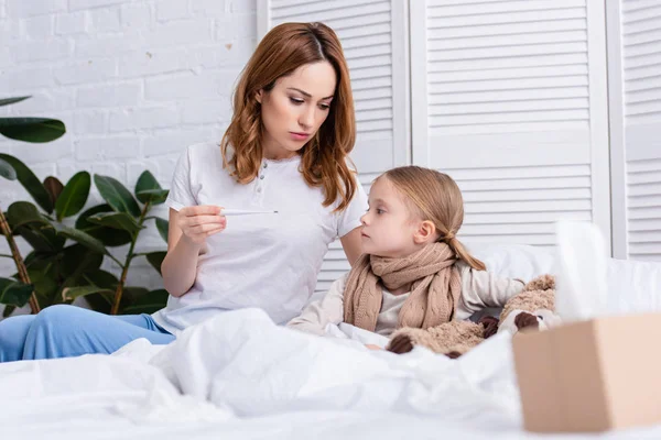 Surface level of mother taking care of sick daughter in bedroom and checking her temperature with thermometer — Stock Photo