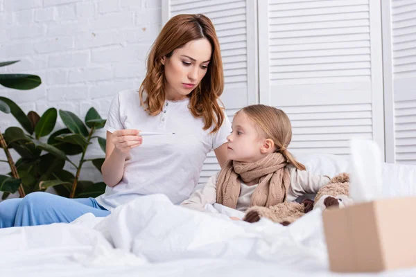 Nível de superfície da mãe cuidando da filha doente no quarto e verificando sua temperatura com termômetro — Fotografia de Stock