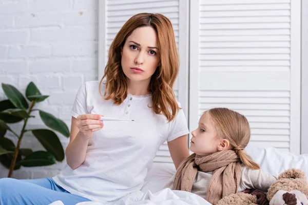 Madre cuidando a la hija enferma en el dormitorio y comprobando su temperatura con termómetro, mirando a la cámara - foto de stock