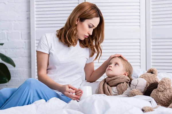 Madre cuidando de la hija enferma en el dormitorio y dándole una taza de té, mirándose - foto de stock