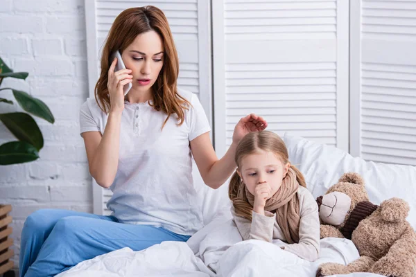 Mother calling doctor by smartphone and palming sick daughter in bed — Stock Photo