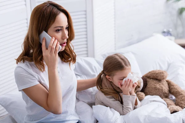 Mãe chamando médico por smartphone, filha doente soprando nariz na cama — Fotografia de Stock