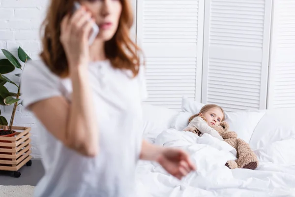 Selective focus of mother calling doctor by smartphone, sick daughter in bed — Stock Photo