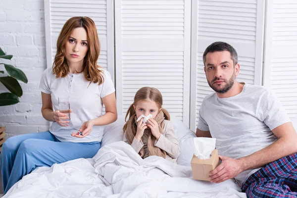 Eltern kümmern sich um kranke Tochter im Schlafzimmer, geben ihr Tabletten und ein Glas Wasser, schauen in die Kamera — Stockfoto