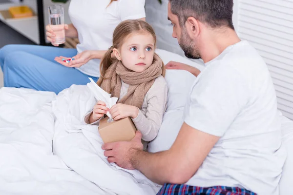 Parents taking care of sick daughter in bedroom, giving her pills, glass of water and napkins — Stock Photo