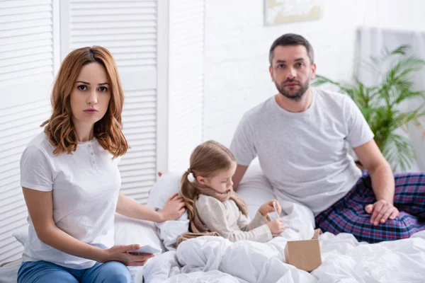 Besorgte Eltern sitzen neben kranker Tochter im Schlafzimmer und schauen in die Kamera — Stockfoto