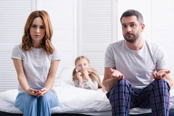 Padres preocupados sentados cerca de hija enferma en el dormitorio y mirando a la cámara, padre mostrando gesto encogiéndose de hombros - foto de stock