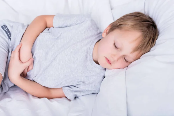 Niño enfermo acostado en la cama y tocando el estómago doloroso en el dormitorio - foto de stock