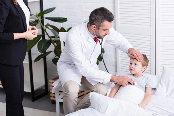 Pediatra de bata blanca examinando niño enfermo con estetoscopio, madre con chaqueta esperando en la habitación - foto de stock