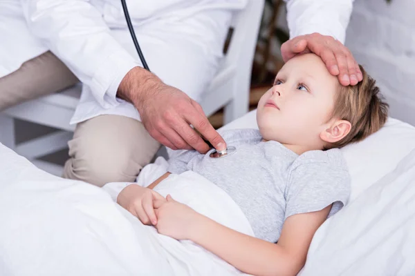 Imagen recortada de pediatra de capa blanca examinando niño enfermo con estetoscopio y tocando la frente - foto de stock