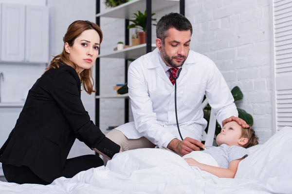Pediatra de bata blanca examinando al niño enfermo con estetoscopio, madre con chaqueta sentada en la cama - foto de stock
