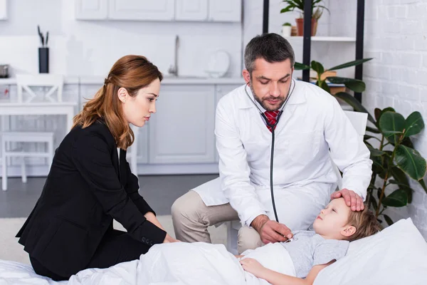 Pediatra bonito em casaco branco examinando menino doente com estetoscópio, mãe de jaqueta sentada na cama — Fotografia de Stock
