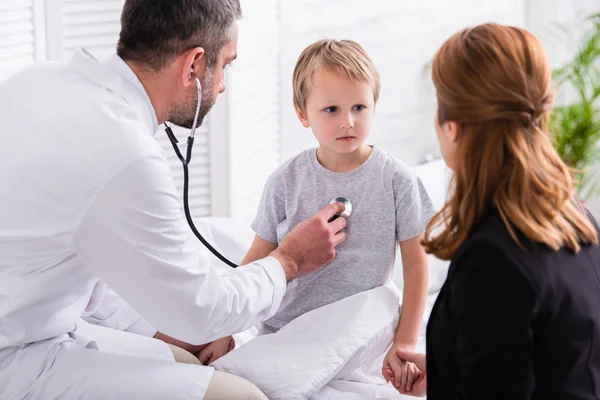Pediatrist in white coat examining sick boy with stethoscope at home — Stock Photo