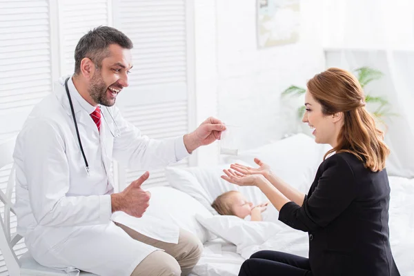 Happy pediatrist in white coat giving medicines to mother of sick boy at home and showing thumb up — Stock Photo