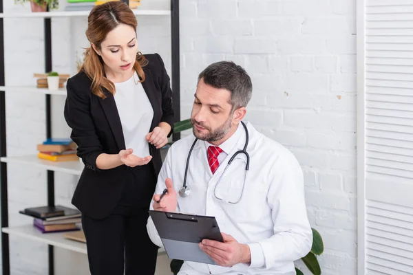 Doctor in white coat writing something to clipboard to woman in suit at home — Stock Photo