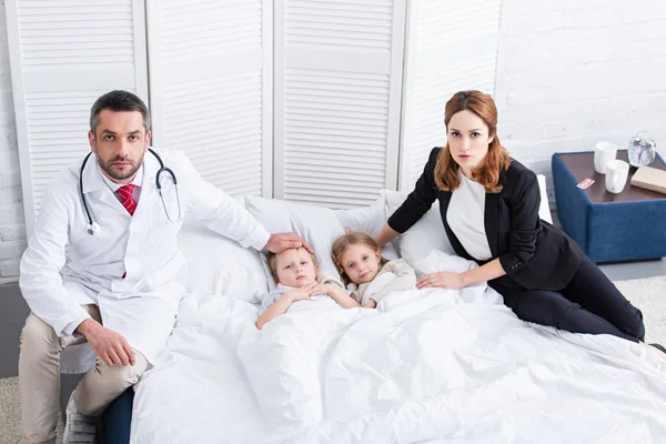 Pédiatre et mère assise sur le lit avec deux enfants malades et regardant la caméra dans la chambre — Photo de stock