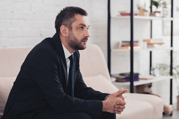Side view of worried handsome businessman in suit sitting on sofa in office and looking away — Stock Photo