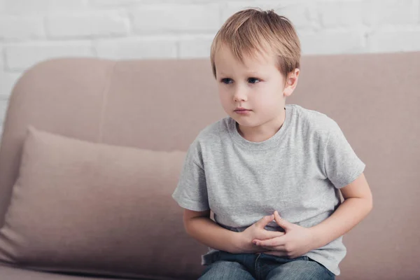 Niño enfermo con dolor de estómago sentado en el sofá en la sala de estar y mirando hacia otro lado - foto de stock