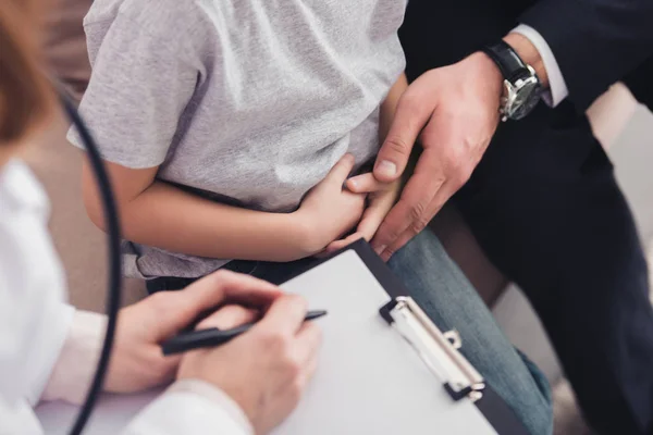 Cropped image of pediatrist writing something in clipboard about sick boy in living room — Stock Photo