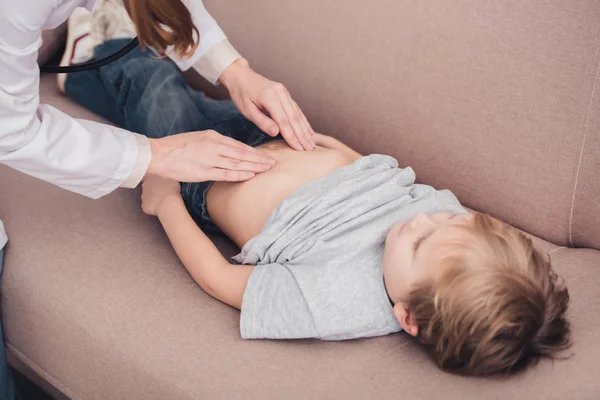 Imagen recortada de pediatra en pelaje blanco palpando estómago de niño enfermo en sofá en sala de estar - foto de stock