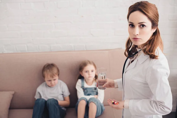 Pediatra sosteniendo pastillas y vaso de agua para niños enfermos en la sala de estar - foto de stock