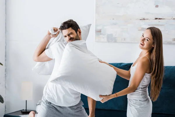 Attractive emotional woman hitting boyfriend with pillow — Stock Photo