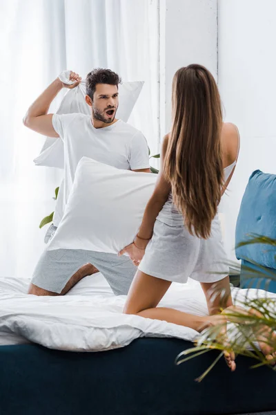 Hombre guapo con expresión de cara divertida teniendo pelea de almohadas con la novia - foto de stock