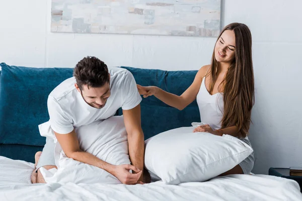 Mujer atractiva joven sentada en la cama con almohada blanca y novio conmovedor - foto de stock