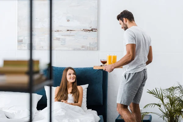 Selective focus of young man holding wooden tray with breakfast while woman lying in bed — Stock Photo