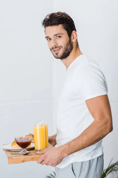 Handsome man holding wooden tray with breakfast and looking at camera — Stock Photo