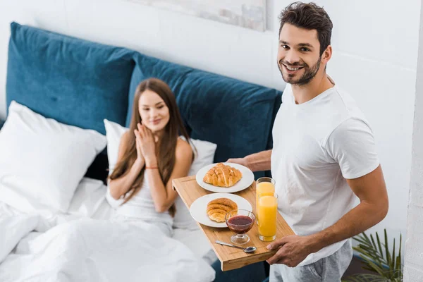 Smiling man holding wooden tray with breakfast while girl sitting in bed — Stock Photo