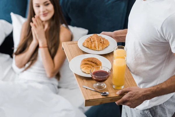 Cropped view of man holding wooden tray with breakfast while girl sitting in bed — Stock Photo