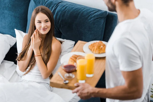 Joven mujer sonriente mirando novio con desayuno en bandeja de madera - foto de stock